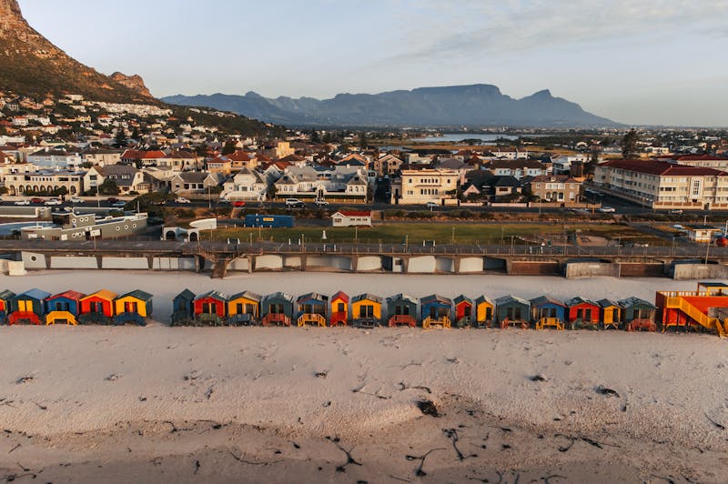 A row of colorful houses along a white-sand beach with houses and mountains as backdrops in Muizenberg Cape Town